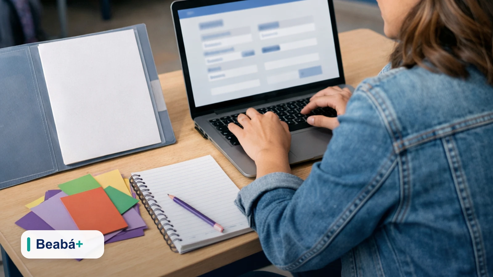Professora cadastrando e configurando o perfil de um aluno no notebook, em uma mesa com materiais escolares.